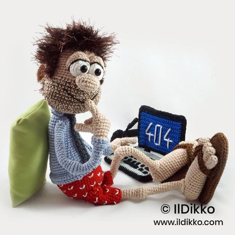 May include: A crocheted doll with messy brown hair, a light blue shirt, and red shorts with white heart patterns. The doll sits with a finger to its chin, facing a black crocheted laptop displaying the text 404 in white on a blue screen.