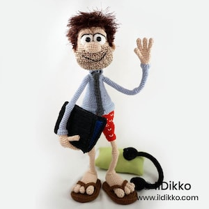 May include: A crocheted doll with brown hair, a light blue shirt, a dark tie, and red patterned shorts. The doll holds a black rectangular object, wears brown sandals, and has a small green pillow and a black corded accessory nearby. Text reads Ildikko.