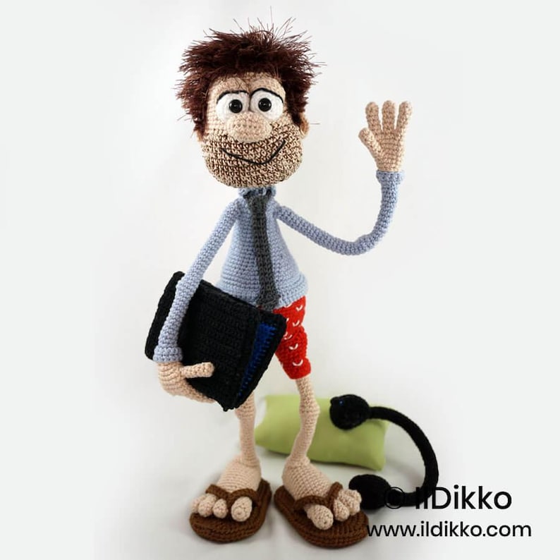 May include: A crocheted doll with brown hair, a light blue shirt, a dark tie, and red patterned shorts. The doll holds a black rectangular object, wears brown sandals, and has a small green pillow and a black corded accessory nearby. Text reads Ildikko.