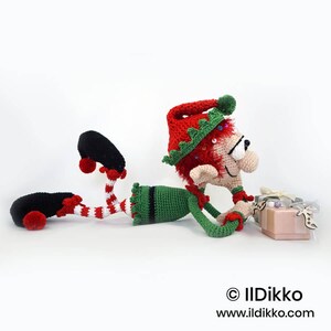May include: A crocheted elf doll with red hair, wearing a red and green hat, a green tunic, and red and white striped leggings with black shoes. The elf is lying down next to two small gift boxes. Text reads: &copy; IlDikko www.ildikko.com.