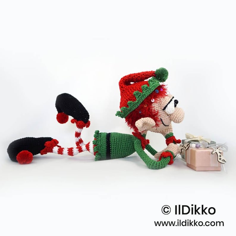May include: A crocheted elf doll with red hair, wearing a red and green hat, a green tunic, and red and white striped leggings with black shoes. The elf is lying down next to two small gift boxes. Text reads: &copy; IlDikko www.ildikko.com.