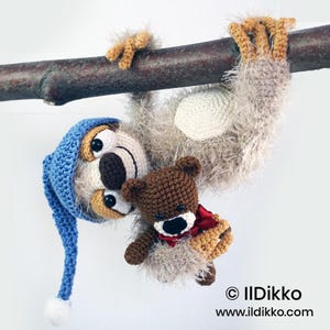 Peut inclure: Un paresseux au crochet en fil beige duveteux, portant un bonnet bleu pointu avec un pompon blanc, suspendu à une branche. Le paresseux tient un petit ours en peluche marron au crochet avec un nœud papillon rouge. Texte en bas : © IlDikko www.ildikko.com