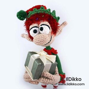 May include: A crocheted elf doll with bright red hair, large white eyes, and pointed ears, wearing a green and red outfit with a matching hat. The elf holds a square gift box wrapped in sage green patterned paper with a cream-coloured ribbon.