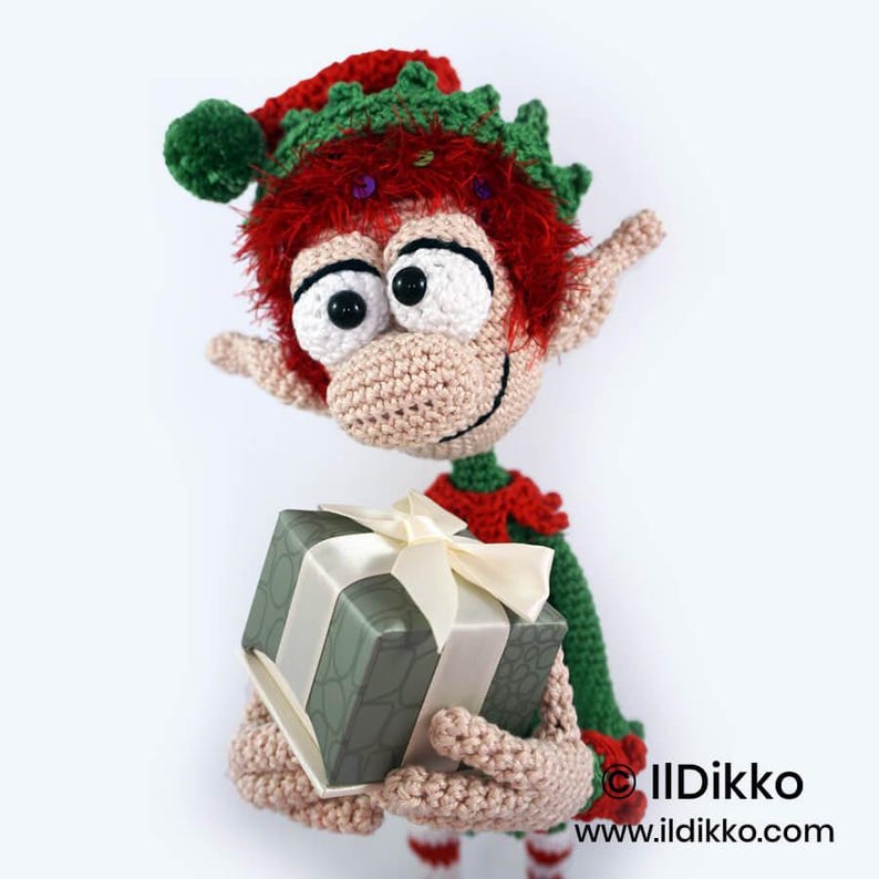 May include: A crocheted elf doll with bright red hair, large white eyes, and pointed ears, wearing a green and red outfit with a matching hat. The elf holds a square gift box wrapped in sage green patterned paper with a cream-coloured ribbon.