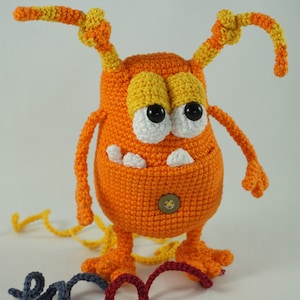 Puede incluir: Juguete monstruo de crochet naranja con detalles de hilo amarillo y naranja. El monstruo tiene ojos grandes blancos con pupilas negras, una nariz de botón y una sonrisa de dientes. El juguete está sentado sobre una superficie blanca con una cola de hilo rojo y gris.