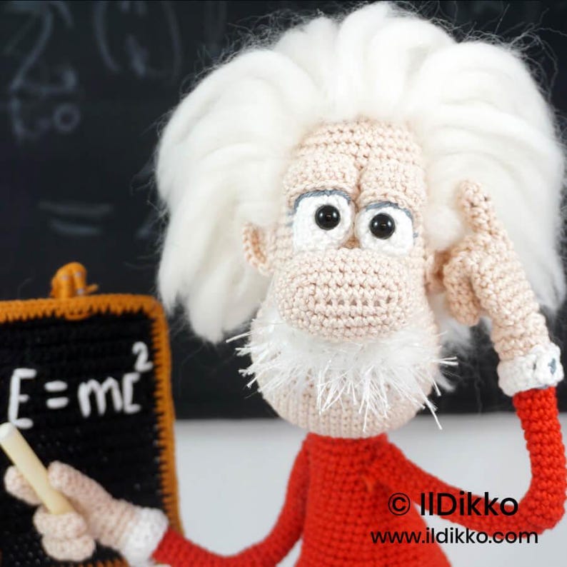 May include: A crocheted doll of a scientist with wild white hair, a white beard, and a red long-sleeved shirt. The doll holds a small black chalkboard with the white text E=mc2 and a piece of chalk, with one hand raised to its head.