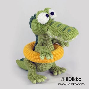 May include: A green crocheted crocodile toy with large white eyes and dark pupils, wearing a bright yellow crocheted swim ring around its midsection. The text "&copy; Ildikko www.ildikko.com" is printed in black in the bottom right corner.