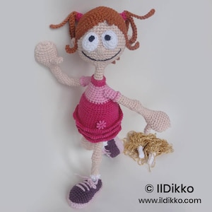 May include: A crocheted doll with light brown yarn hair in pigtails, wearing a pink dress with a small flower detail and dark purple sneakers. The doll has large white eyes and holds a gold-colored pom-pom. Text reads: © Ildikko www.ildikko.com