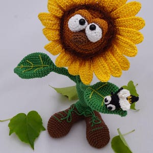 May include: A crocheted sunflower with a brown face, large white eyes, and a black nose. The sunflower has green leaves and brown boots. A small crocheted bee is perched on one of the leaves. The sunflower is standing on a white surface with green leaves scattered around it. The image includes the text "&copy; Ildikko www.ildikko-crochet.com".