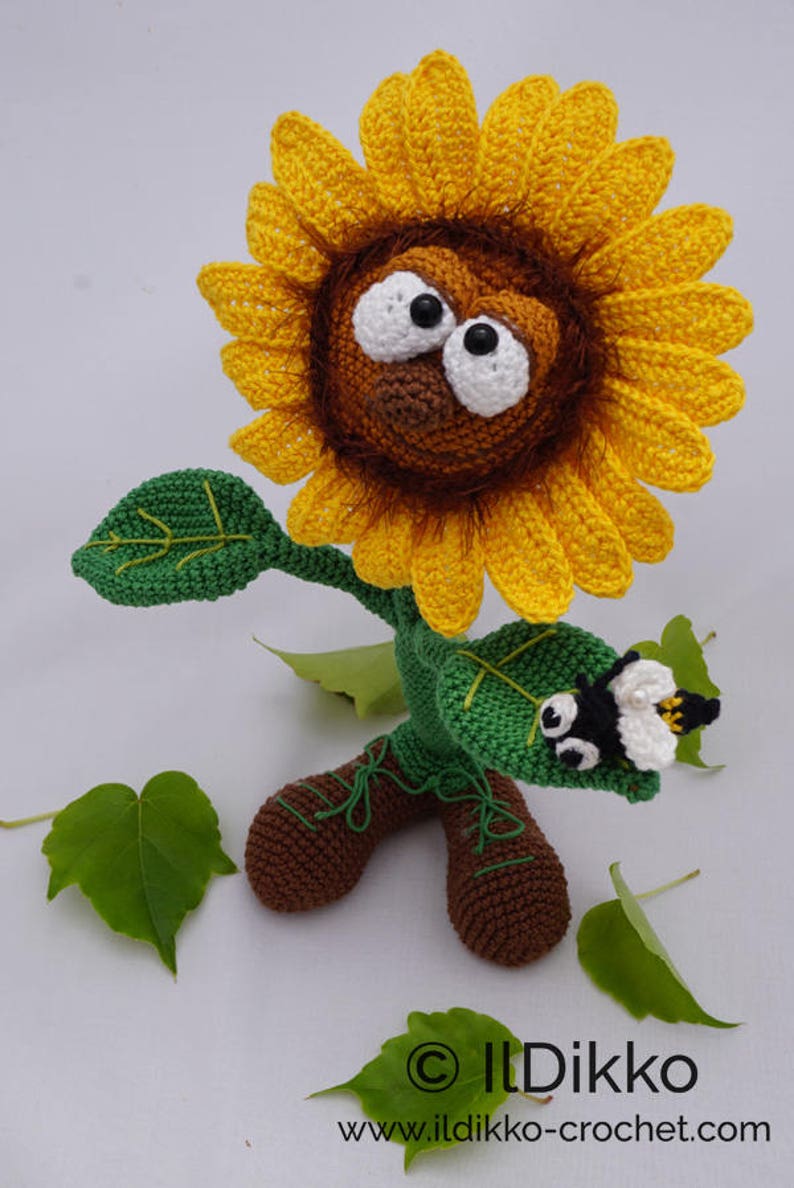 May include: A crocheted sunflower with a brown face, large white eyes, and a black nose. The sunflower has green leaves and brown boots. A small crocheted bee is perched on one of the leaves. The sunflower is standing on a white surface with green leaves scattered around it. The image includes the text "&copy; Ildikko www.ildikko-crochet.com".