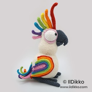May include: A handmade crochet parrot toy in cream, featuring a rainbow crest, tail, and wing accents. The bird has a large, round eye with a pink outline and a dark gray beak. The parrot is sitting upright.