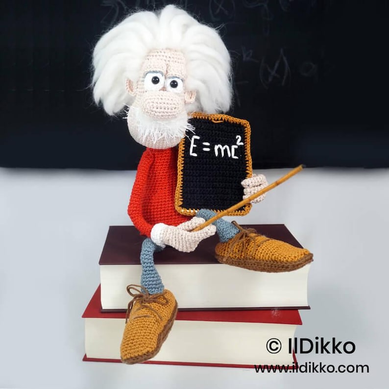 May include: A crocheted doll with wild white hair, a red long-sleeved shirt, and blue trousers, sitting on two stacked books. The doll holds a black rectangular chalkboard with the white text E=mc2 and a thin wooden pointer stick.