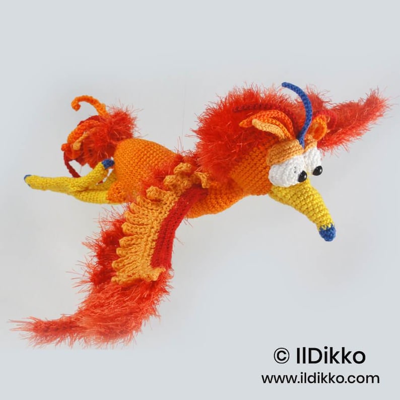 May include: A crocheted phoenix toy featuring vibrant orange and red yarn with fuzzy textures on the wings and head. It has large white eyes with black pupils, a yellow beak with a blue tip, and a blue curved crest. Text reads: &copy; IlDikko www.ildikko.com