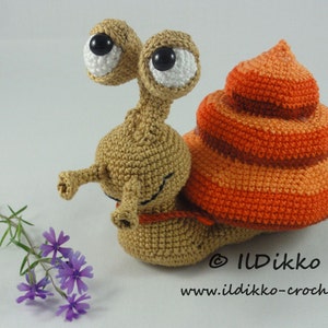 May include: A crocheted snail toy with large eyes, a brown body, and an orange and red shell. The snail is sitting on a white surface with a small sprig of purple flowers.