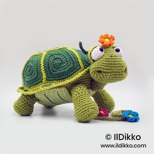 May include: A crocheted turtle toy featuring a green body, a shell with dark green spiral patterns and yellow borders, and large white eyes. An orange flower sits on its head, and a string with small colorful flowers trails from its side.