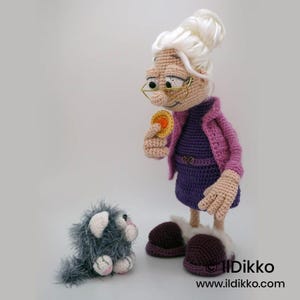 May include: A crocheted doll of an elderly woman wearing glasses, a purple dress, and a pink jacket, holding a yellow coin. A small, gray crocheted cat sits nearby. The doll has white hair and brown shoes. The doll is handmade.