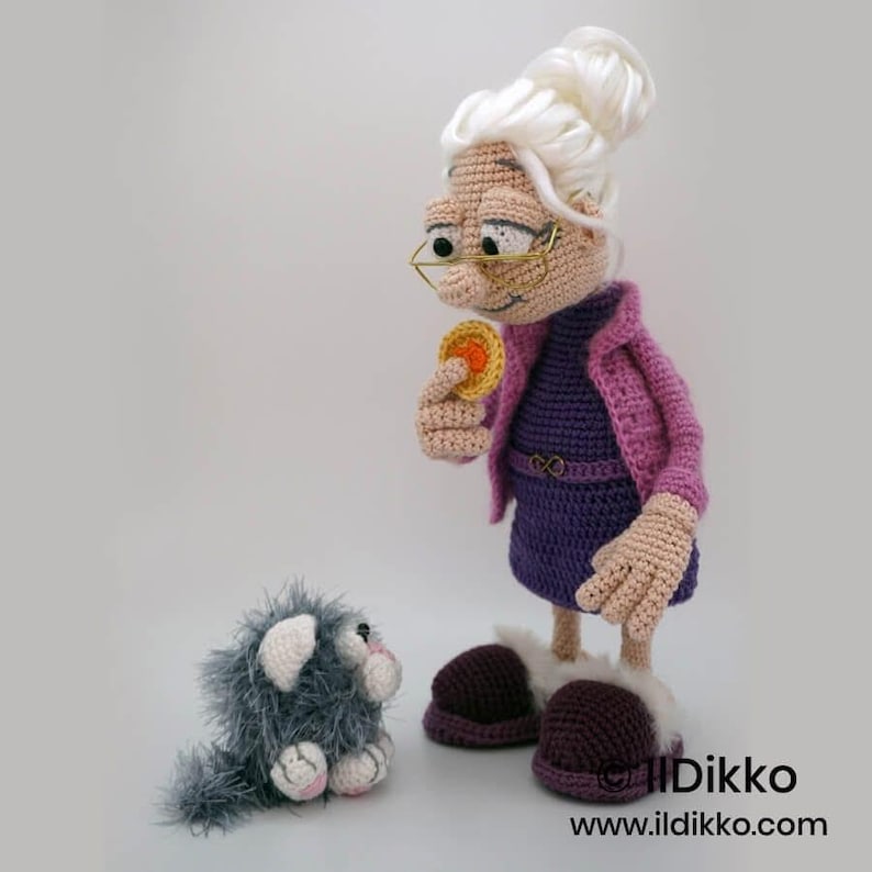 May include: A crocheted doll of an elderly woman wearing glasses, a purple dress, and a pink jacket, holding a yellow coin. A small, gray crocheted cat sits nearby. The doll has white hair and brown shoes. The doll is handmade.