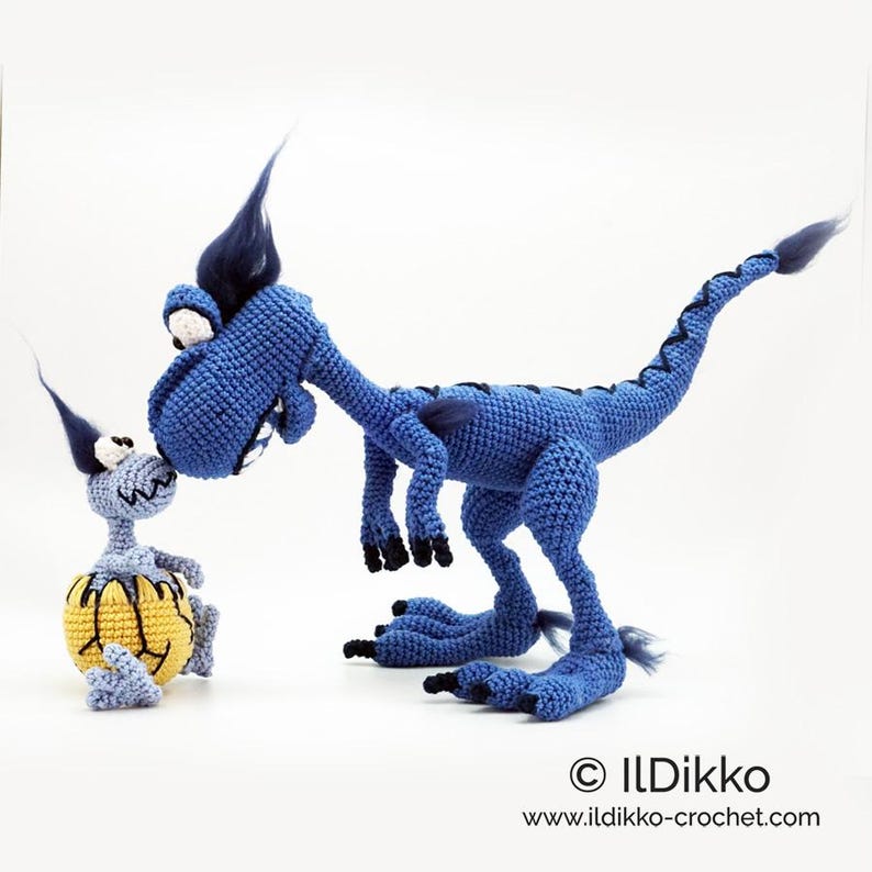 May include: A large blue crocheted dinosaur with dark blue hair and black claws standing next to a smaller light blue crocheted creature emerging from a yellow and black egg. The text ILDikko and www.ildikko-crochet.com is visible.