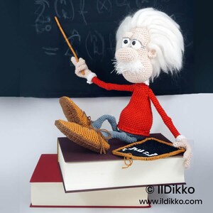 May include: A crocheted doll of an elderly scientist with wild white hair, a mustache, and glasses, wearing a red long-sleeved shirt and gray pants. The doll sits on two stacked books, holding a thin orange pointer and a small black chalkboard.