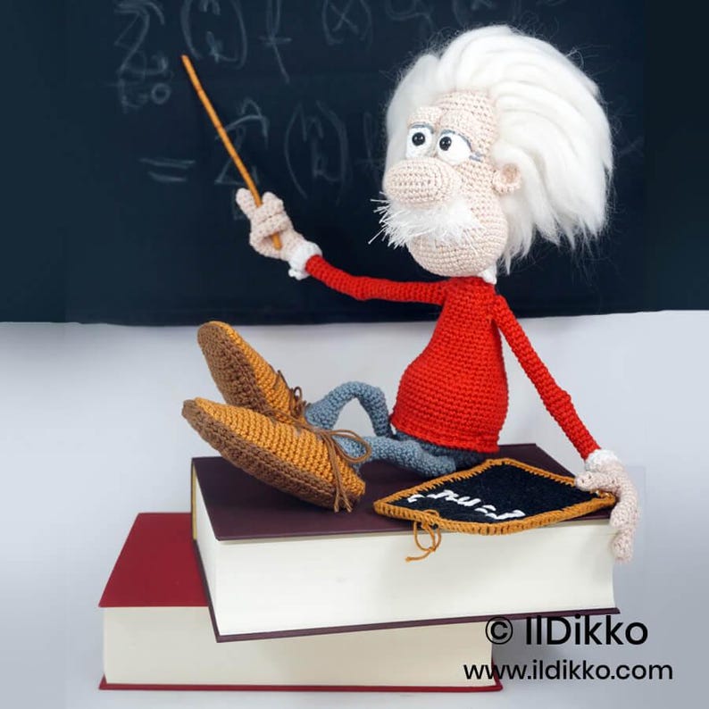 May include: A crocheted doll of an elderly scientist with wild white hair, a mustache, and glasses, wearing a red long-sleeved shirt and gray pants. The doll sits on two stacked books, holding a thin orange pointer and a small black chalkboard.