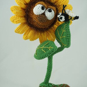 May include: A crocheted sunflower with a brown face and large eyes, wearing brown shoes. A crocheted bee is perched on a green leaf of the sunflower.