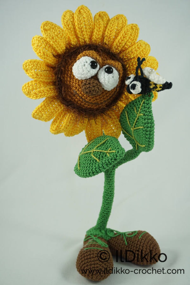 May include: A crocheted sunflower with a brown face and large eyes, wearing brown shoes. A crocheted bee is perched on a green leaf of the sunflower.
