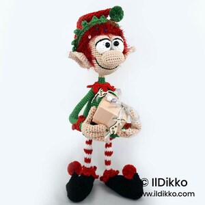 May include: A handcrafted crochet elf doll with red hair, wearing a green and red outfit with a matching hat. The elf has large white eyes, pointed ears, and striped legs. It holds a small beige gift box decorated with a silver ribbon and a wooden charm.
