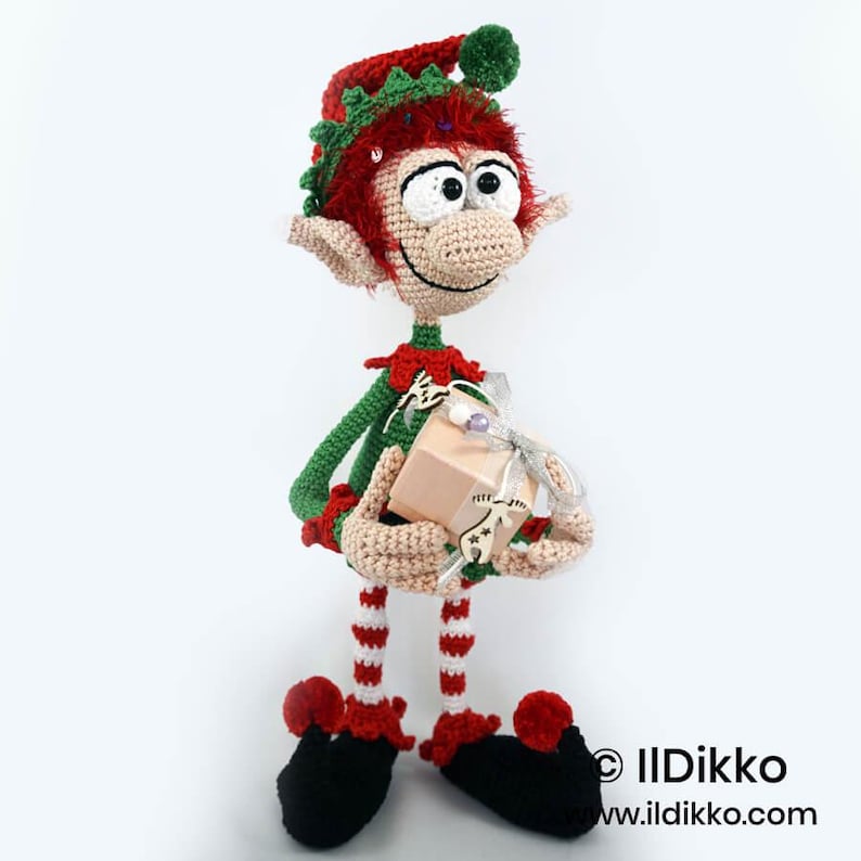 May include: A handcrafted crochet elf doll with red hair, wearing a green and red outfit with a matching hat. The elf has large white eyes, pointed ears, and striped legs. It holds a small beige gift box decorated with a silver ribbon and a wooden charm.