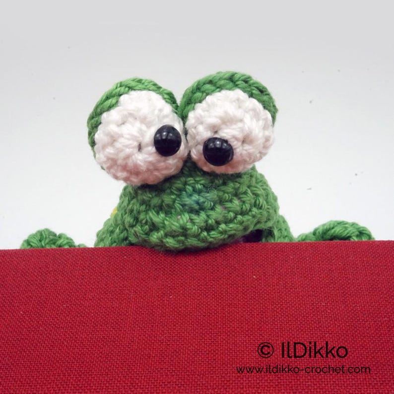 May include: A green crocheted frog with large white eyes and black pupils peeking over the top of a red surface. The frog is a handmade amigurumi toy.