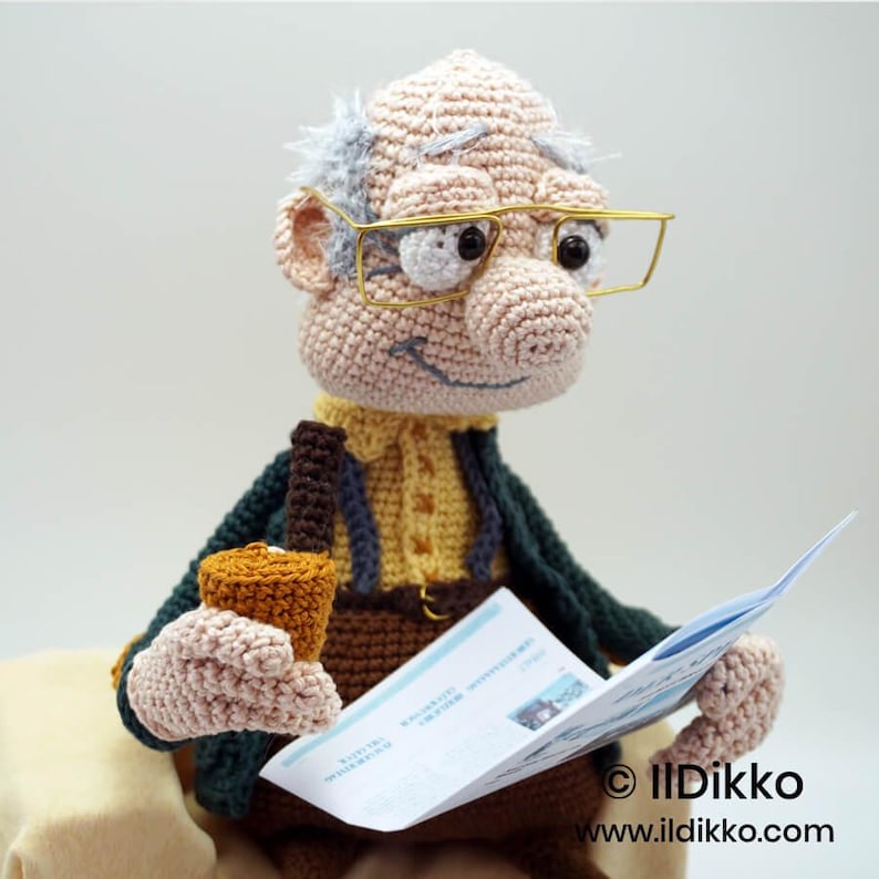 May include: A crocheted doll of an elderly man wearing gold-rimmed glasses, a yellow shirt, and a dark green jacket. The doll is holding a small brown cup and a folded paper. The doll has gray hair and is smiling. The doll is handmade.