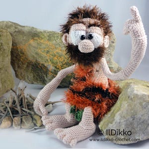May include: A crocheted caveman doll with light skin, a dark brown fuzzy beard and hair, and large white eyes with black pupils. The doll wears a textured orange and black garment with a small green leaf detail and has one arm raised.