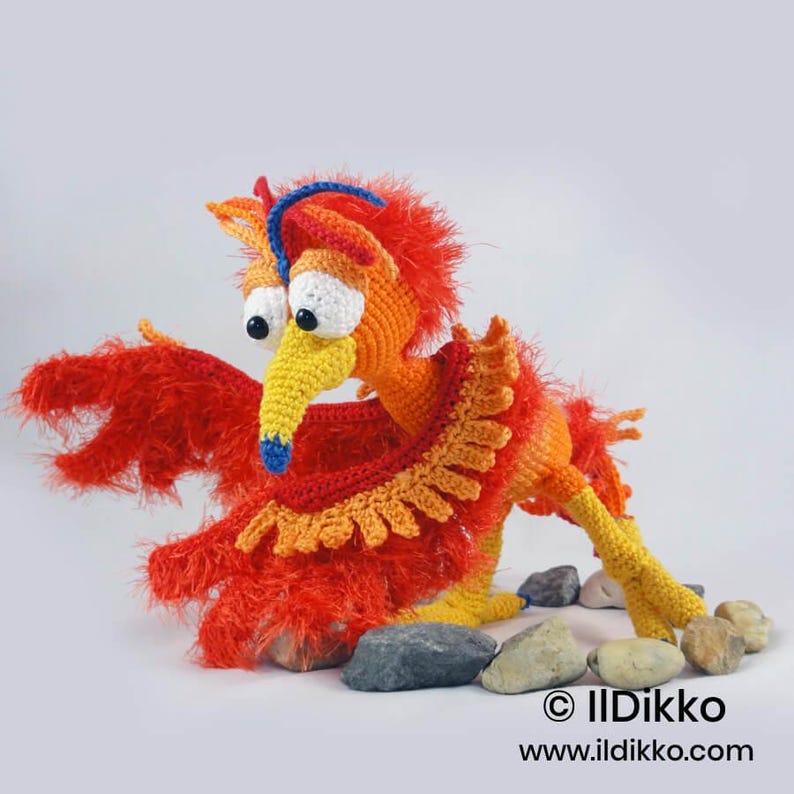 May include: A crocheted phoenix toy featuring bright orange and red fuzzy yarn, a yellow beak with a blue tip, and large white eyes with black pupils. The creature has a blue crest and a ruffled collar, positioned among several smooth gray stones.