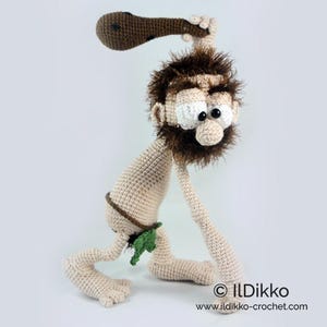 May include: A crocheted caveman doll with beige skin, a brown beard, and messy hair. The figure wears a green leaf loincloth and holds a brown club with black spots overhead. The text ILDikko and www.ildikko-crochet.com is printed at the bottom.