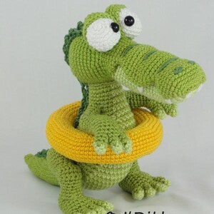 May include: A green crocheted crocodile toy with large white eyes and black pupils, wearing a bright yellow crocheted swim ring around its midsection. The crocodile has a textured back and webbed feet. Text reads ILDikko and www.ildikko-crochet.com.
