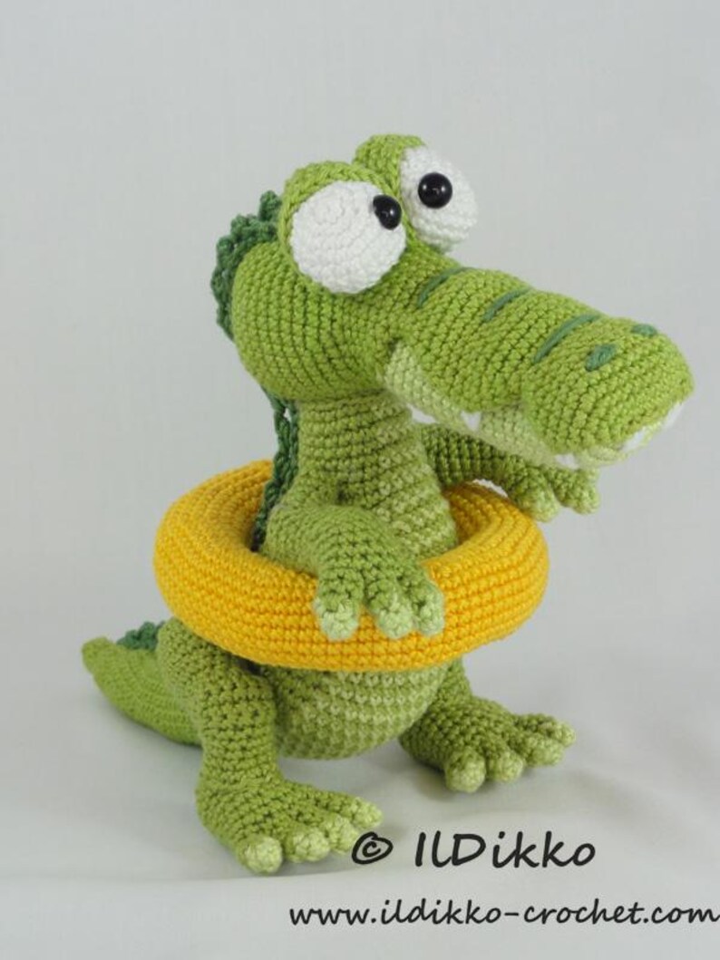 May include: A green crocheted crocodile toy with large white eyes and black pupils, wearing a bright yellow crocheted swim ring around its midsection. The crocodile has a textured back and webbed feet. Text reads ILDikko and www.ildikko-crochet.com.