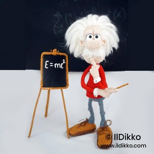 May include: A crocheted doll of a scientist with wild white hair, a red long-sleeved shirt, and gray trousers. The doll wears brown shoes and stands next to a small easel holding a black board with the white text E=mc2 written on it.