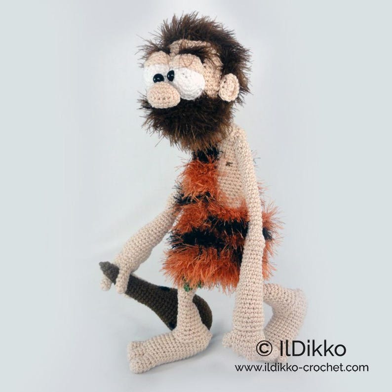 May include: A crocheted caveman doll with beige skin, a shaggy dark brown beard and hair, and large white eyes with black pupils. The figure wears a fuzzy orange and black striped tunic and holds a dark brown crocheted club in one hand.