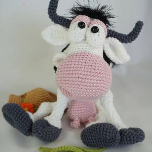 Peut inclure: Une vache en crochet rose et blanche avec des cornes et des cils noirs. La vache est assise sur une surface blanche avec un chapeau en crochet marron et une fleur en crochet verte.