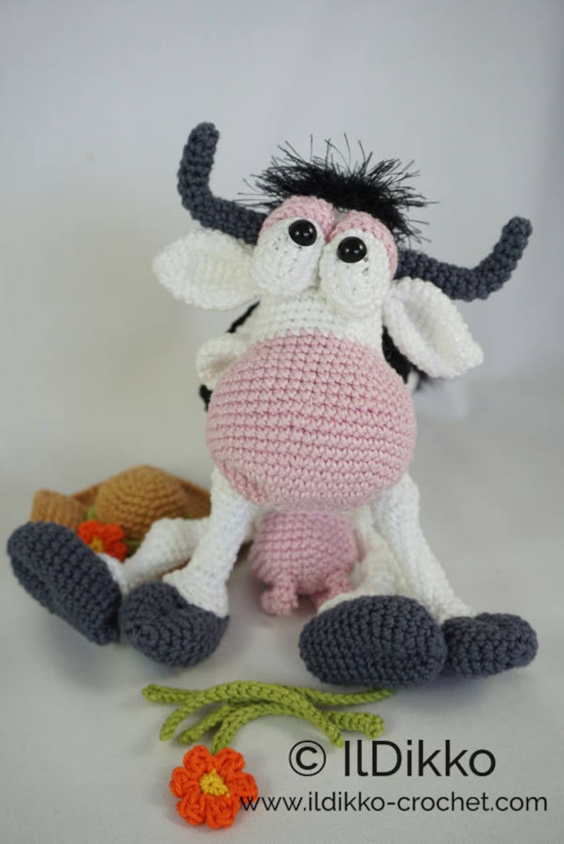 Peut inclure: Une vache en crochet rose et blanche avec des cornes et des cils noirs. La vache est assise sur une surface blanche avec un chapeau en crochet marron et une fleur en crochet verte.