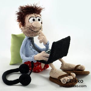 May include: A crocheted doll with messy brown hair, a light blue button-down shirt, and red patterned shorts. The doll has a beard, large white eyes, and is sitting while using a black laptop. A pair of black headphones and brown sandals are visible.