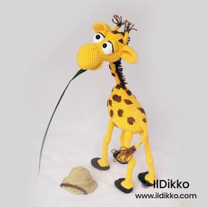 May include: A yellow and brown crocheted giraffe toy with large white eyes, a long neck, and dark brown hooves. The giraffe holds a long green leaf in its mouth. A small, tan crocheted safari hat sits on the ground next to the giraffe figure.