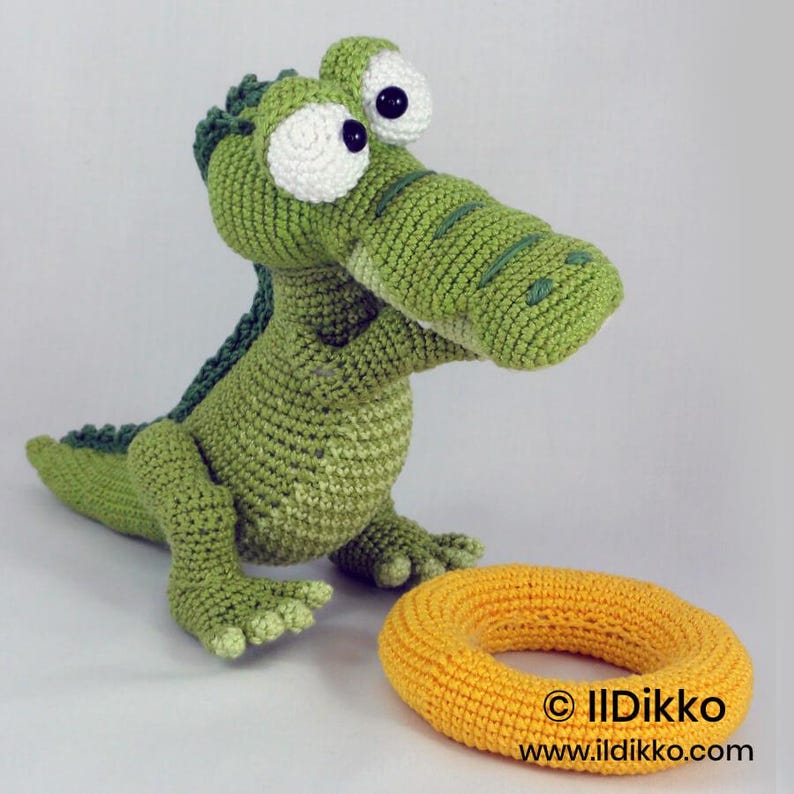 May include: A green crocheted crocodile toy with large white eyes and dark green back scales, positioned next to a yellow crocheted ring. The text "&copy; IlDikko www.ildikko.com" is printed in black near the bottom right of the image.