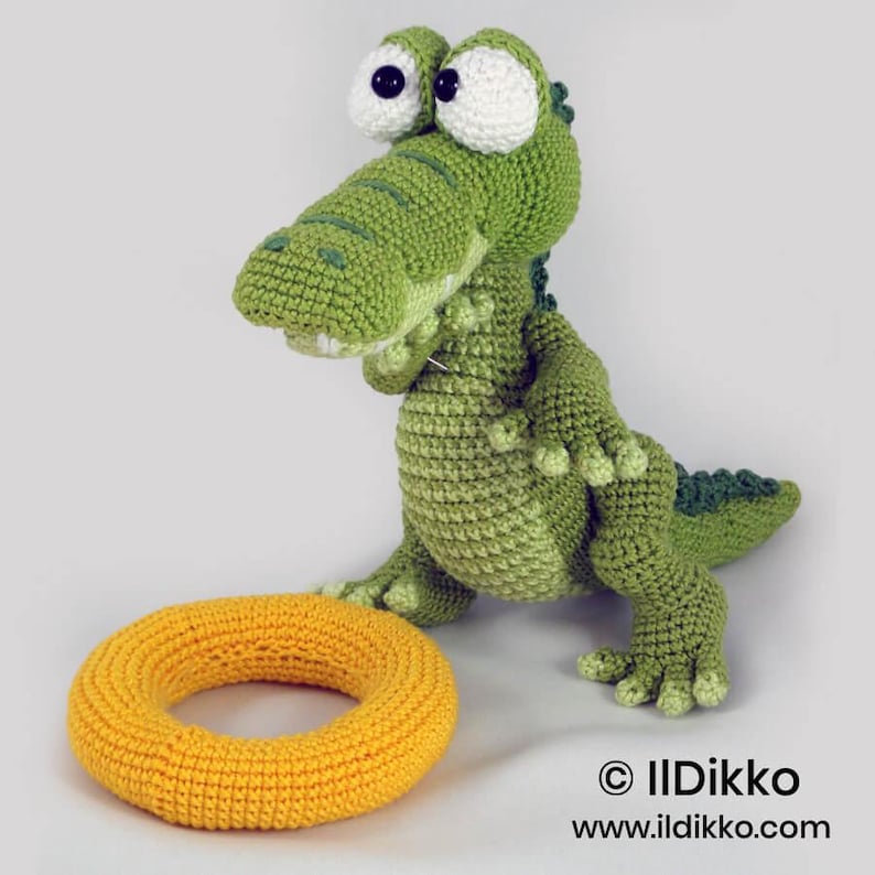 May include: A green crocheted crocodile toy with large white eyes and dark pupils, standing next to a yellow crocheted ring. The text "&copy; IIDikko www.ildikko.com" is printed in the bottom right corner of the image.