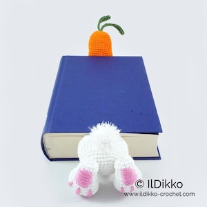 May include: A white crocheted bunny bookmark with pink feet peeking out from behind a blue book. The bunny is holding an orange crocheted carrot.