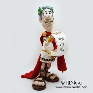 Op de afbeelding: Gehaakte Romeinse soldatenpop die een rol met de tekst "veni vidi vici" vasthoudt. De pop draagt een witte tuniek met een gouden riem, een rode cape en bruine sandalen. De pop heeft groen haar en een bruine baard.