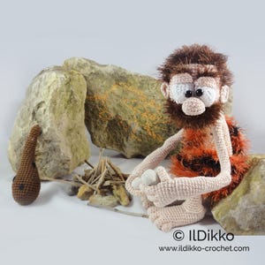 May include: A crocheted caveman doll with brown hair and beard, wearing an orange and black animal print garment. The doll sits near a small pile of sticks and a brown crocheted club. Text at the bottom reads ILDikko www.ildikko-crochet.com.
