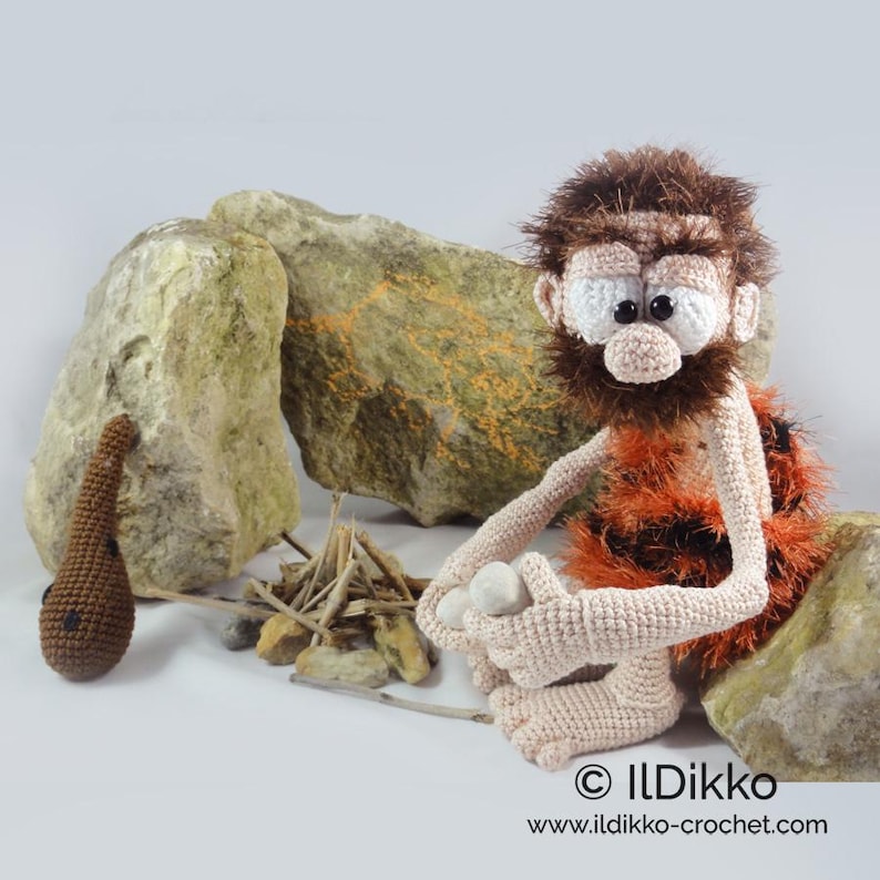 May include: A crocheted caveman doll with brown hair and beard, wearing an orange and black animal print garment. The doll sits near a small pile of sticks and a brown crocheted club. Text at the bottom reads ILDikko www.ildikko-crochet.com.