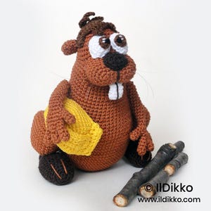 May include: A brown crocheted beaver toy with white eyes, a black nose, and two white teeth. The beaver holds a yellow crocheted log and sits next to two small, real wooden sticks. The text "© Ildikko www.ildikko.com" is visible in the corner.