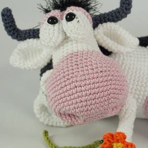 May include: A crocheted cow toy with black and white fur, pink ears, and large black eyes. The cow is lying down with its head turned to the side. It has a green crocheted stem and an orange crocheted flower attached to its leg. The toy is made by Ildikko and the website www.ildikko-crochet.com is displayed on the bottom of the image.