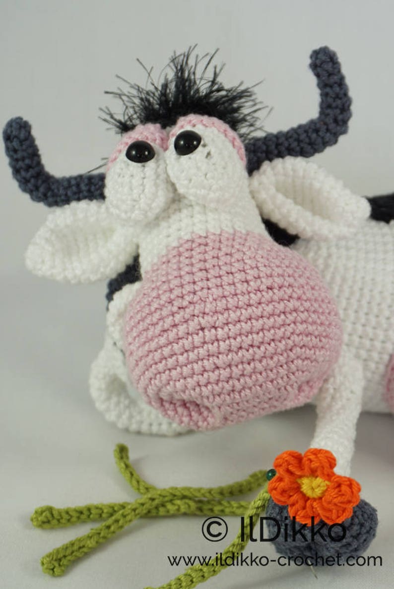 May include: A crocheted cow toy with black and white fur, pink ears, and large black eyes. The cow is lying down with its head turned to the side. It has a green crocheted stem and an orange crocheted flower attached to its leg. The toy is made by Ildikko and the website www.ildikko-crochet.com is displayed on the bottom of the image.